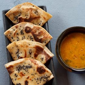 cheese naan wedges served on a black platter with a bowl of curry kept on the right side and text layover.