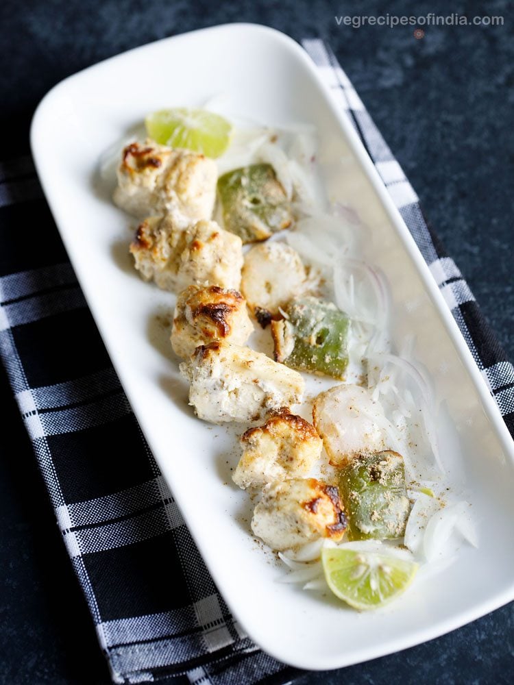 paneer malai tikka served on a rectangular white platter with sliced onion and lemon wedges, and text layover. 