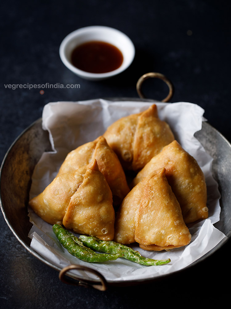 shingara served on a butter paper kept in an iron kadai with fried green chilies, a small bowl of chutney kept on the top side and text layover. 