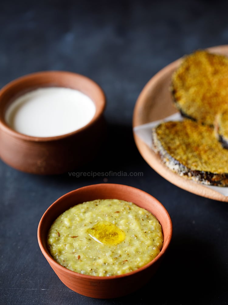 terracotta bowl filled with bajra khichdi topped with melted ghee with a plate of baingan fry and a bowl of buttermilk in the background.