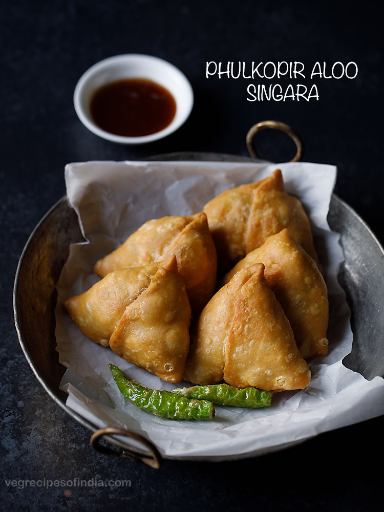 shingara served on a butter paper kept in an iron kadai with fried green chilies, a small bowl of chutney kept on the top side and text layovers. 