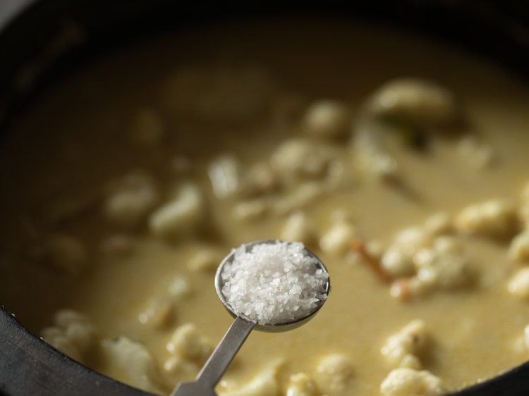 close up of teaspoon of salt held over cauliflower korma cooking in saucepan