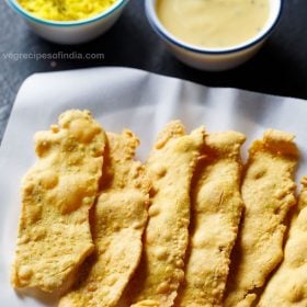 fafda served on a butter paper lined plate with a bowl of papaya sambharo and a bowl of fafda chutney kept on the top in the background with text layover.