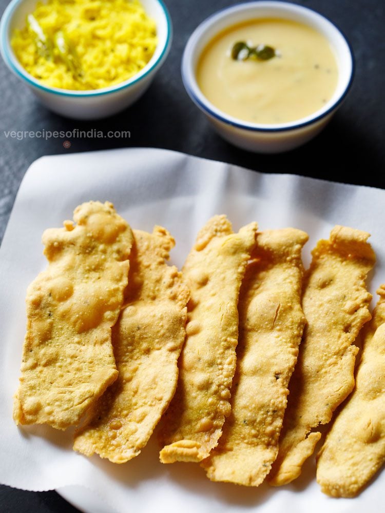 fafda served on a butter paper lined plate with a bowl of papaya sambharo and a bowl of fafda chutney kept on the top in the background with text layover.