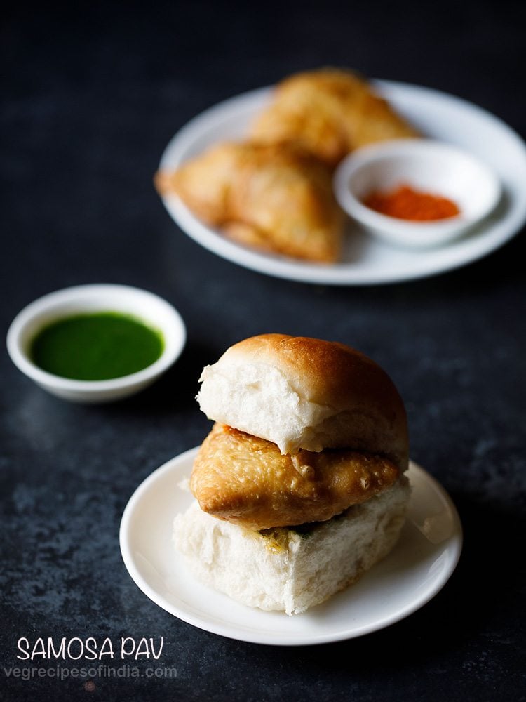 samosa pav served on a white plate with a small bowl of green chutney and a plate of samosas and garlic chutney kept in the background and text layovers. 