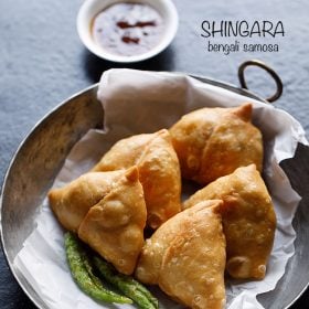 singara served on a butter paper kept in an iron kadai with fried green chilies, a small bowl of chutney kept on the top side and text layovers.