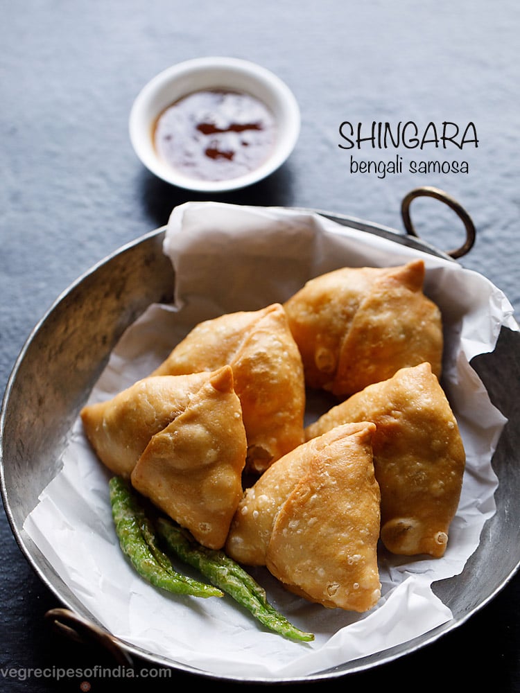 singara served on a butter paper kept in an iron kadai with fried green chilies, a small bowl of chutney kept on the top side and text layovers.