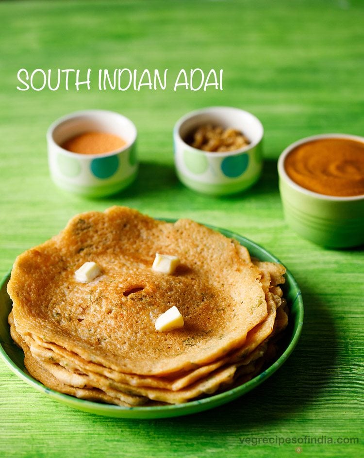 adai stacked and served on a green plate with butter cubes on top and bowls of chutney and sambar kept in the background and text layover. 