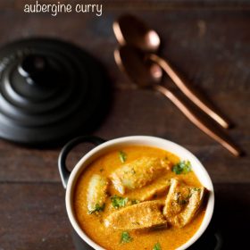 brinjal curry garnished with coriander leaves and served in a black casserole with 2 copper spoons kept in the background and text layover.