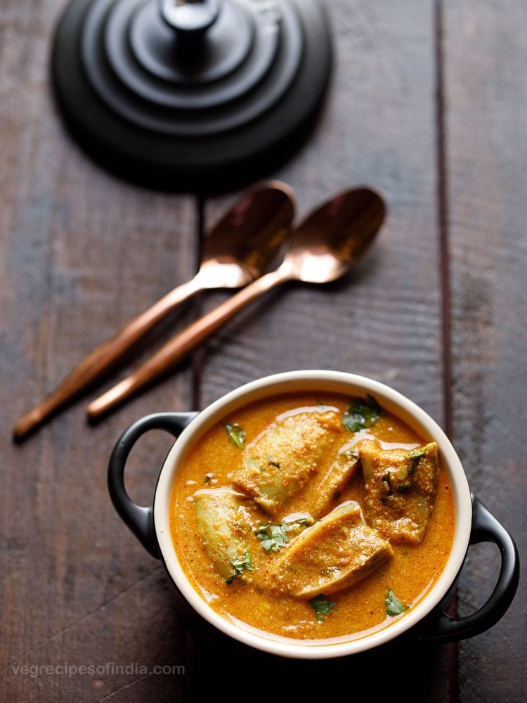 Two copper spoons and black pot lid on wooden table next to bowl of eggplant curry.