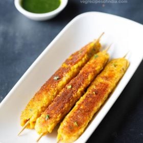 corn kabab skewers served in a white platter with a small bowl of green chutney kept in the top left side and text layover.