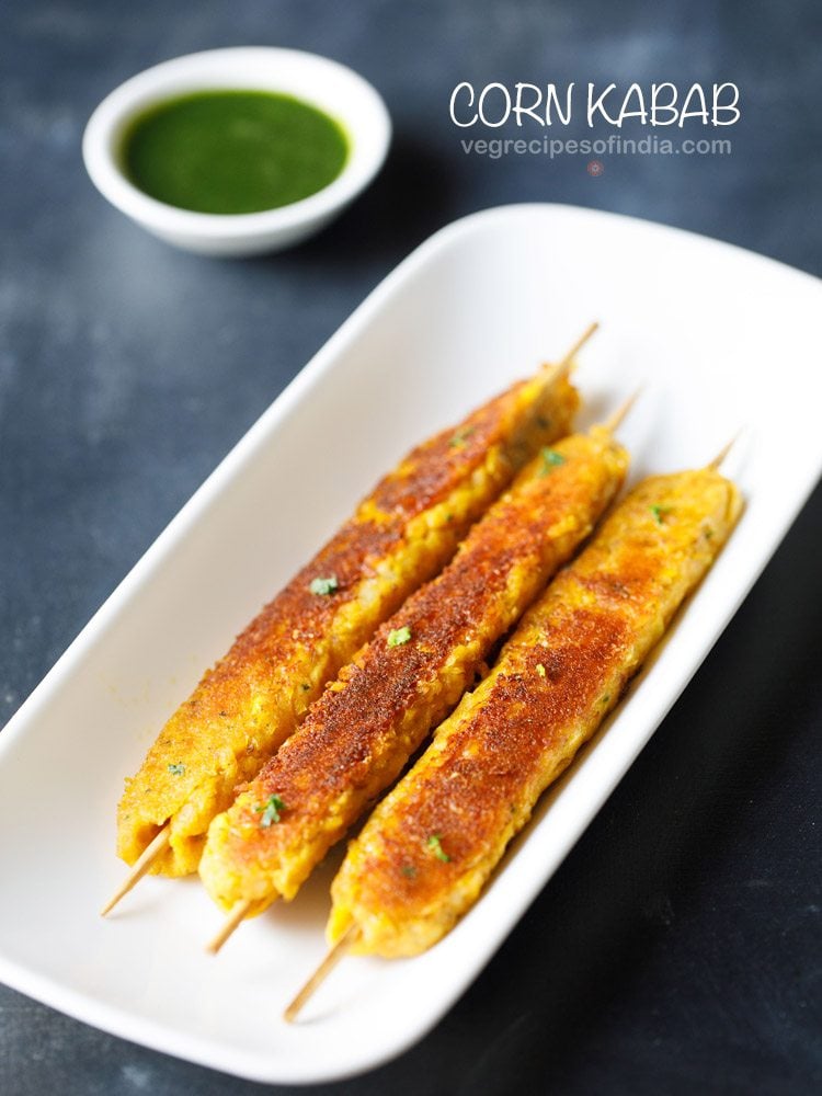 corn kabab skewers served in a white platter with a small bowl of green chutney kept in the top left side and text layover.