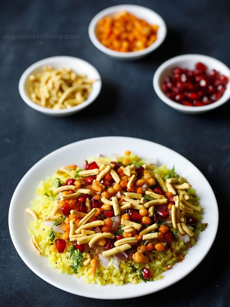 indori poha served on a white plate with small bowls of sev, fresh pomegranate arils and masala boondi kept in the background. 