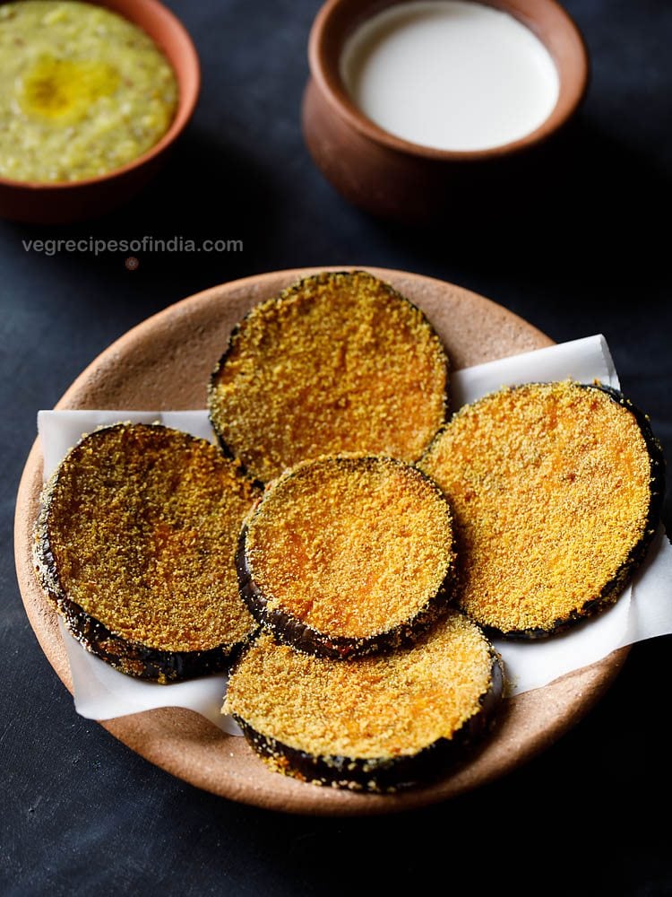 brinjal fry or fried eggplant served on a butter paper placed on a ceramic plate with a bowl of khichdi and curd kept in the background. 
