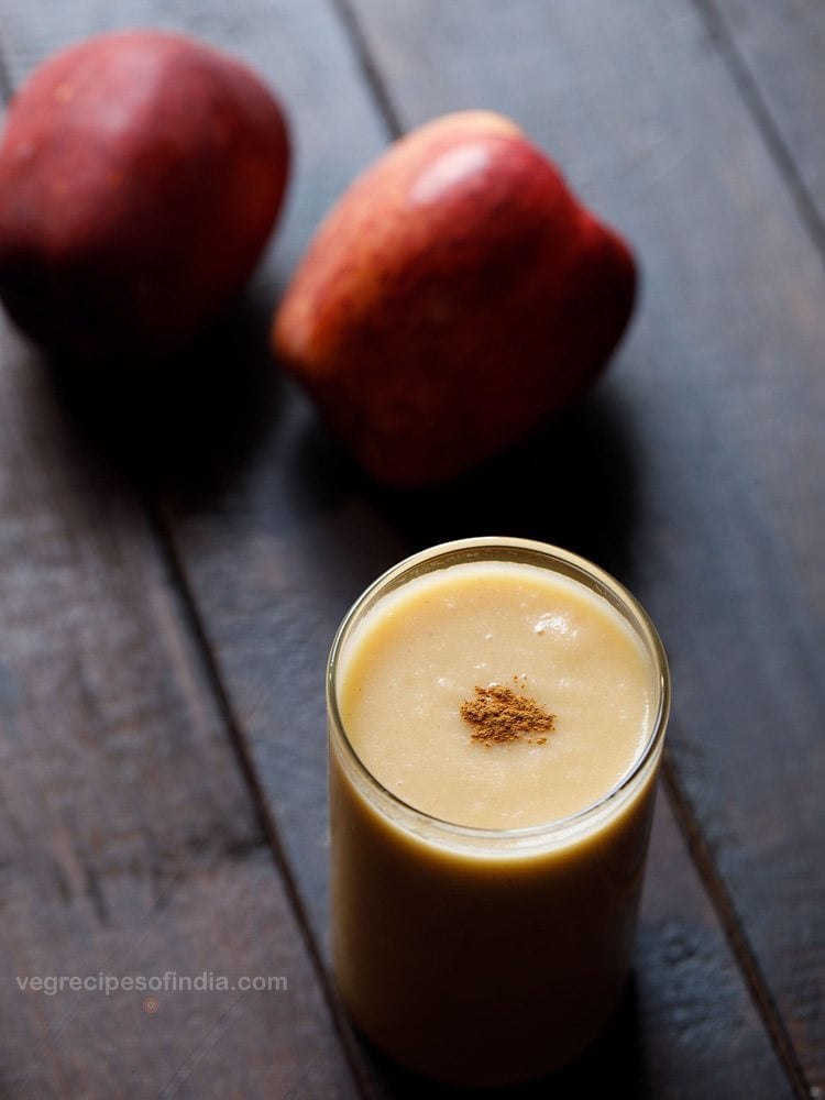 apple smoothie served in a glass garnished with cinnamon powder with two apples kept on the side.  