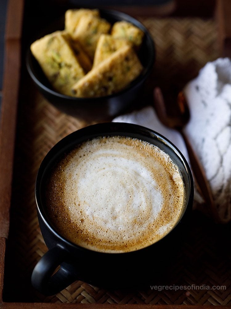 beaten coffee in a black mug on a serving tray with a spoon, napkin, and bowl of treats. 