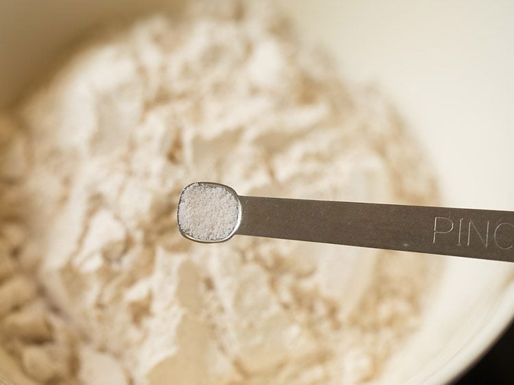 Salt in pinch measurer held above mixing bowl of whole wheat flour.