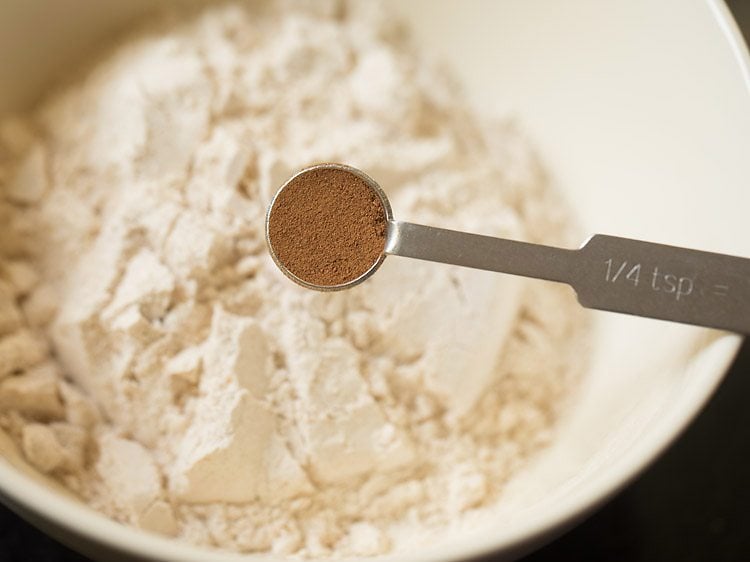 Cinnamon powder in teaspoon measurer held above mixing bowl of whole wheat flour.