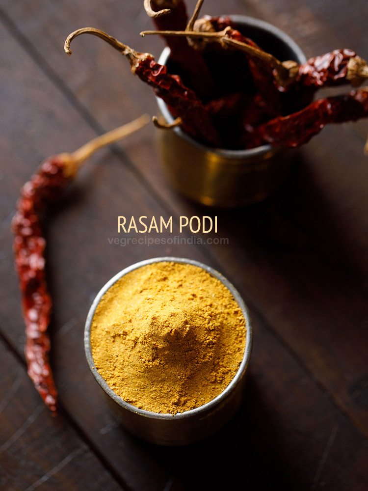 top shot of rasam powder in a brass container with container of dried red chilies in the back and text layover. 