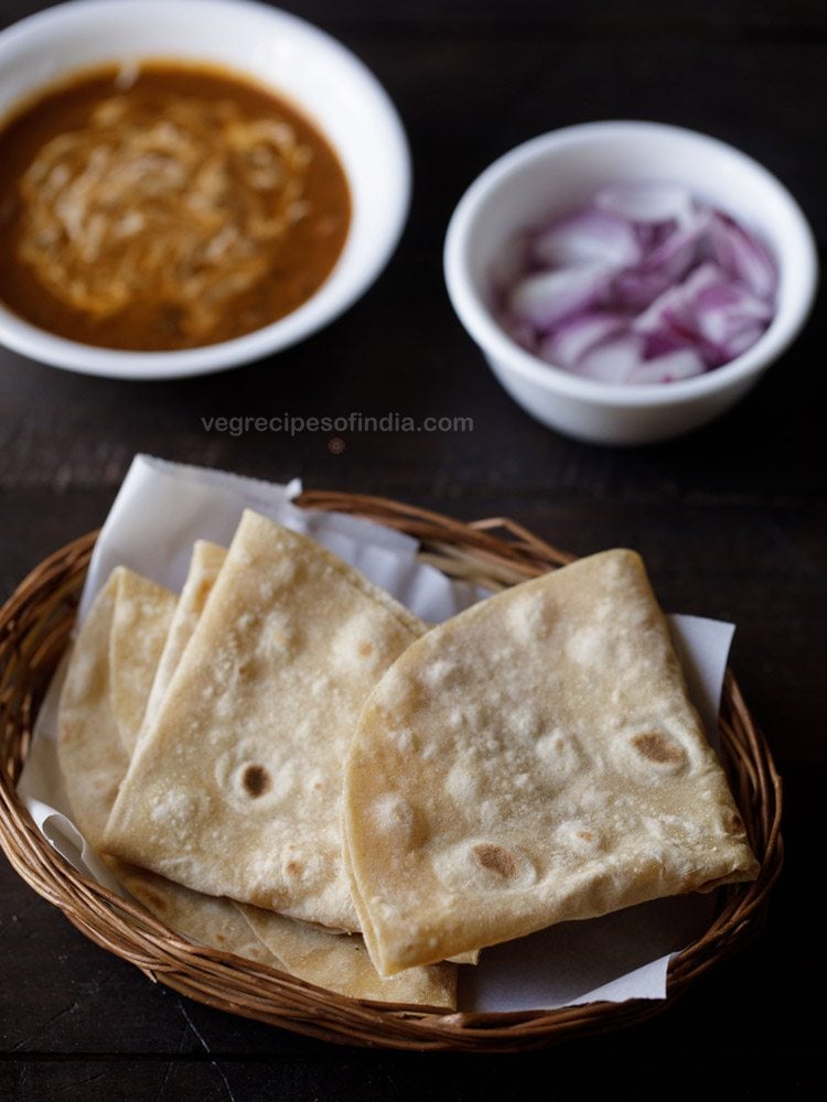 rumali roti folded and served in a basket with a bowl of curry and a bowl of onions kept in the background. 