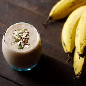 banana smoothie in a glass topped with toasted seeds with a bunch of bananas placed at the side