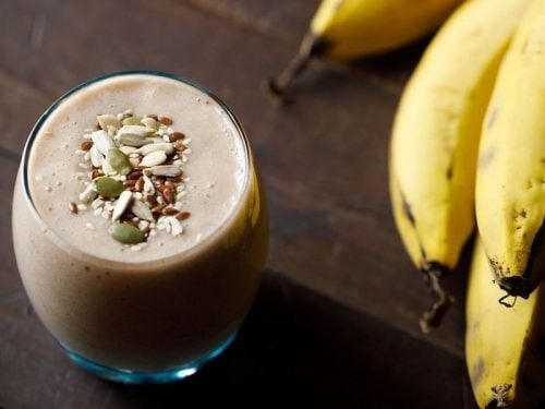 banana smoothie in a glass topped with toasted seeds with a bunch of bananas placed at the side