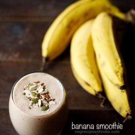 banana smoothie filled in a glass topped with toasted seeds and a bunch of bananas placed at right side on a wooden table