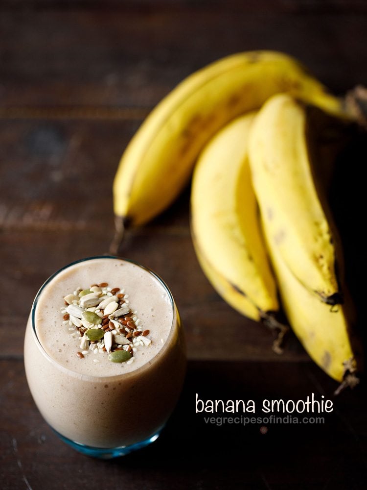 banana smoothie filled in a glass topped with toasted seeds and a bunch of bananas placed at right side on a wooden table