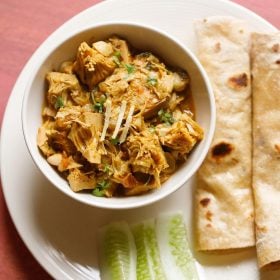 kathal ki sabji served in a white bowl placed on a white plate with rolled chapattis and cucumber fingers kept on it and text layovers.