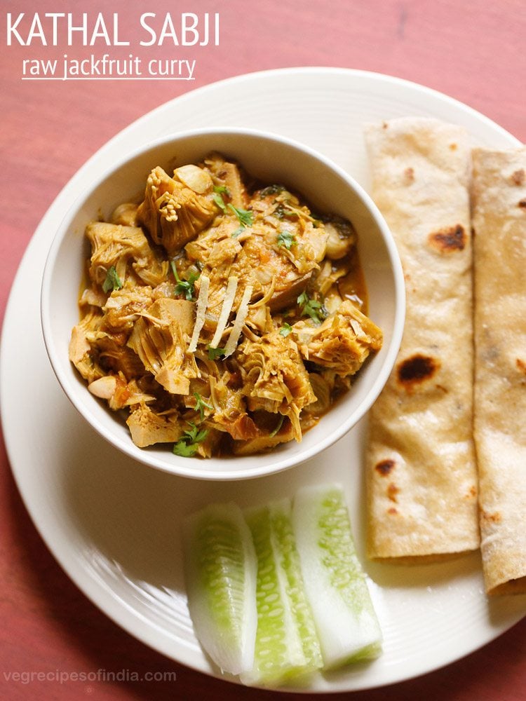 kathal ki sabji served in a white bowl placed on a white plate with rolled chapattis and cucumber fingers kept on it and text layovers.