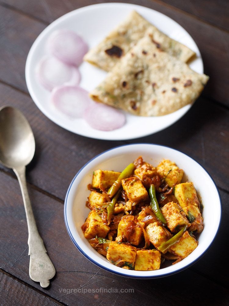paneer fry served in a white bowl with a spoon kept on the left side, chapattis and onion rings kept on a white plate on the top side and text layovers. 