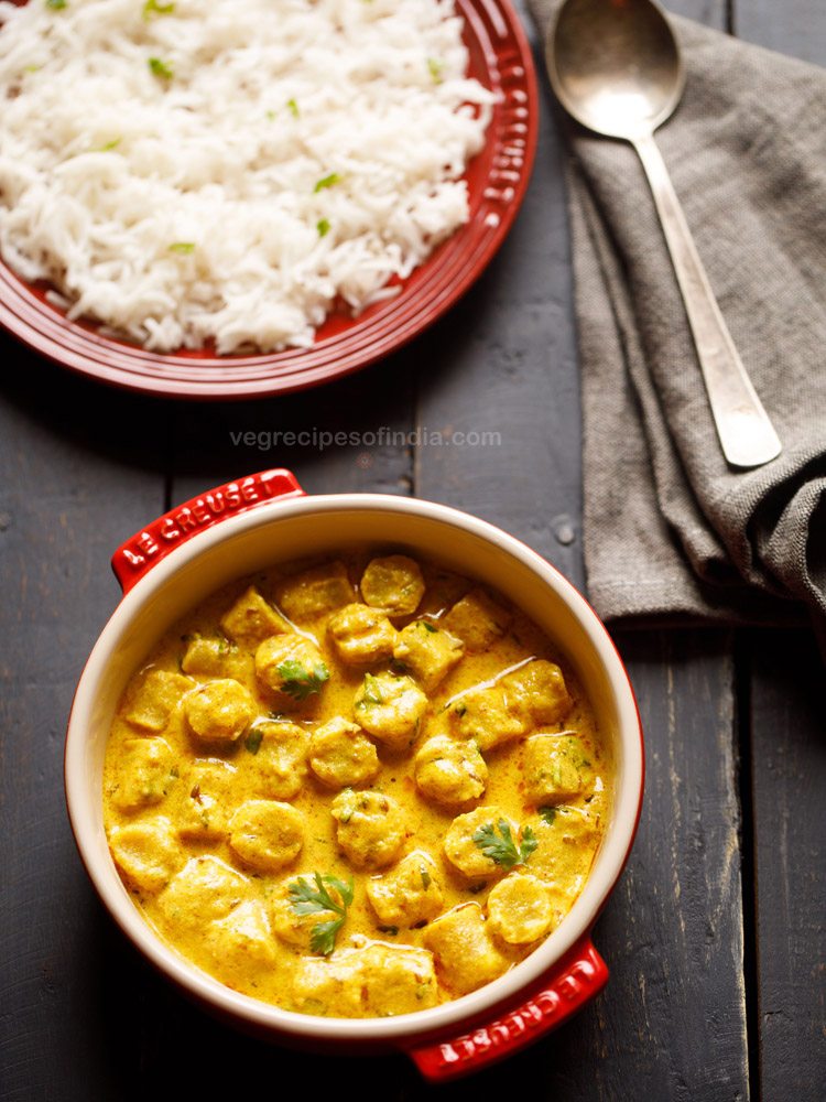 gatte ki sabji garnished with coriander leaves and served in a red casserole with a spoon kept on the right side and a bowl of steamed rice kept on the top left side and text layover. 