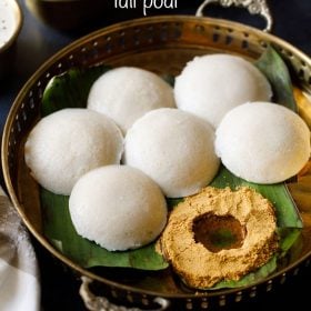 idli podi served with ghee in a well made in it and idlis kept on the side placed on a banana leaf and served on a fancy brass tray with text layover.