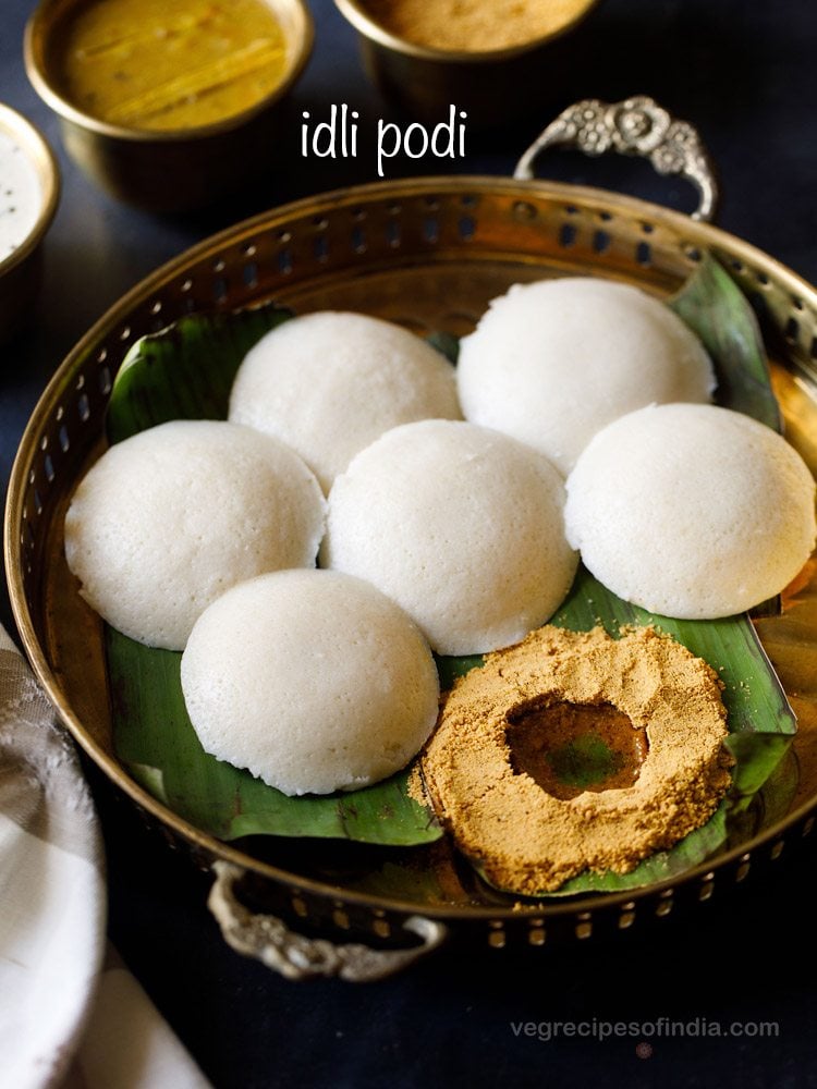 idli podi served with ghee in a well made in it and idlis kept on the side placed on a banana leaf and served on a fancy brass tray with text layover.