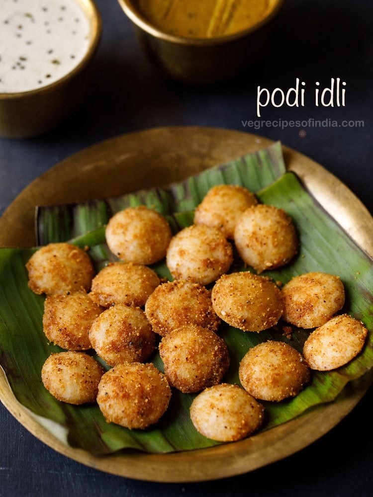 podi idli served on a banana leaf placed on a brass plate with a brass bowl of coconut chutney and brass bowl of sambar kept on the top left side and text layover.