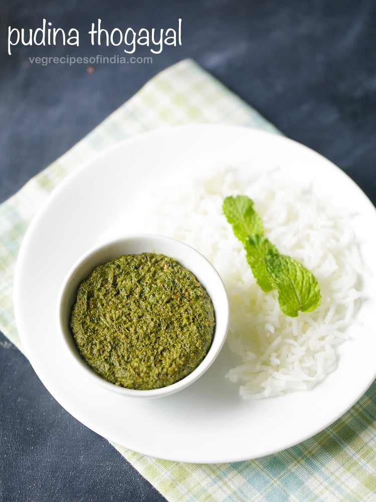 pudina thogayal served in a white bowl placed on white plate with steamed rice garnished with mint leaves and text layover.