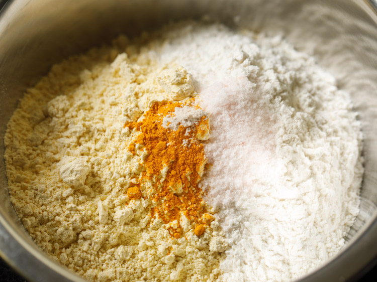 besan, maida, turmeric powder, asafoetida and salt added to a mixing bowl for bhakarwadi recipe. 