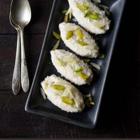 cham cham pieces garnished with sliced pistachios and almonds and served on a black platter with 2 spoons kept on the left side and text layovers.