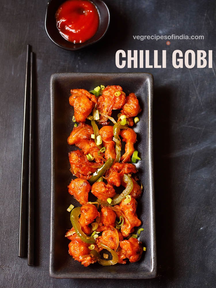 chilli gobi served in a black rectangular tray with black chopsticks on the left side on a blacking grey board with sweet red chilli sauce in a small black bowl placed on top.
