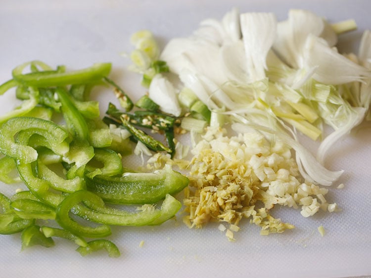 sliced spring onions, capsicum, finely chopped ginger, garlic and slit green chillies on a white chopping board