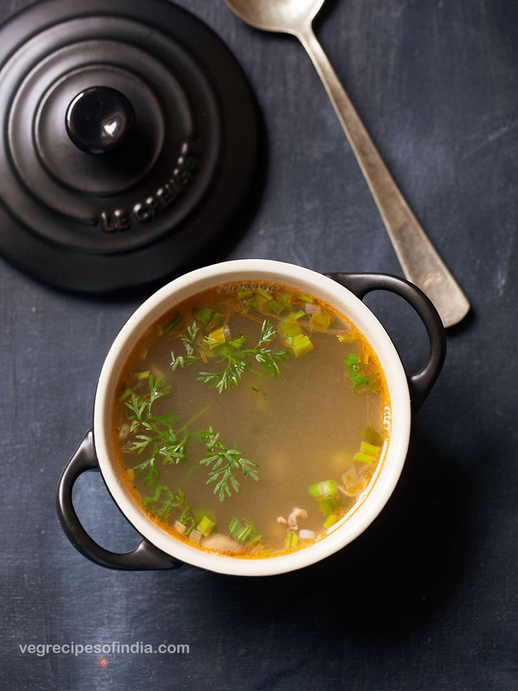 lemon coriander soup served in a black bowl with handles with a soup spoon on the side on a dark bluish grey background.