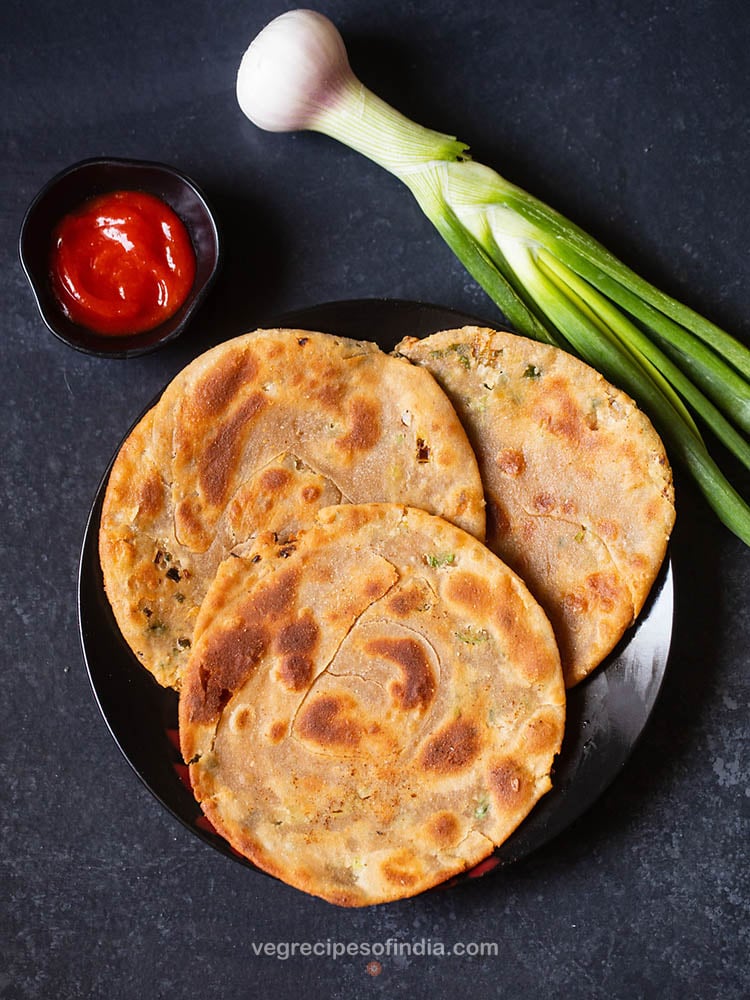 green onion pancakes places on a black ceramic plate on a black board with sriracha sauce served in a small black bowl on left side and a stalk of spring onion placed on the right side of the plate. 