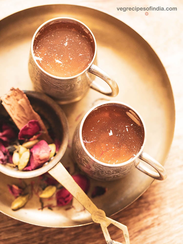 Overhead shot of kahwa tea in an antique silver mug on a copper plate with a small bowl of dried cinnamon bark, cardamom pods and rose petals on the side.