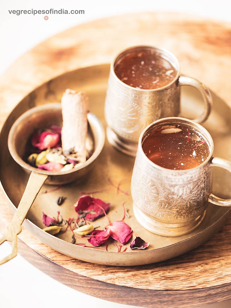 Kashmiri kahwa tea served in 2 small silver cups. a brass strainer filled with green tea leaves, spices, rose petals, saffron kept as the side.