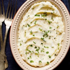 Mashed potatoes in oval ceramic bowl on slate gray wooden board. Garnish with some chopped parsley and rosemary.