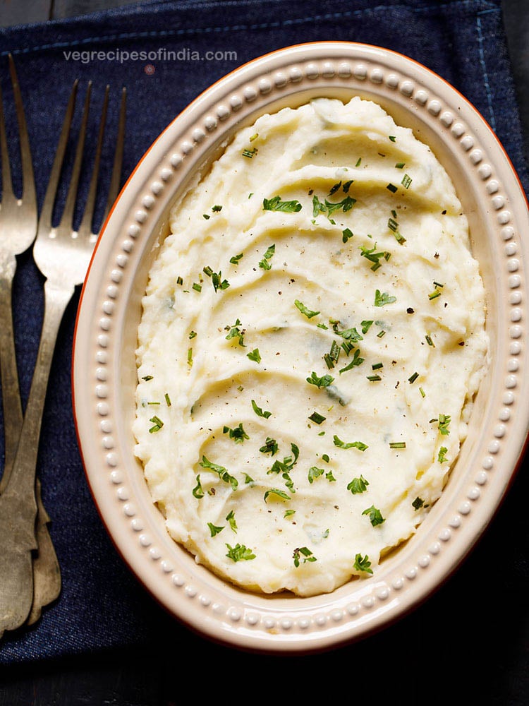 mashed potatoes served in an oval ceramic bowl kept on a slate grey wooden board. garnished with some finely chopped parsley and rosemary.