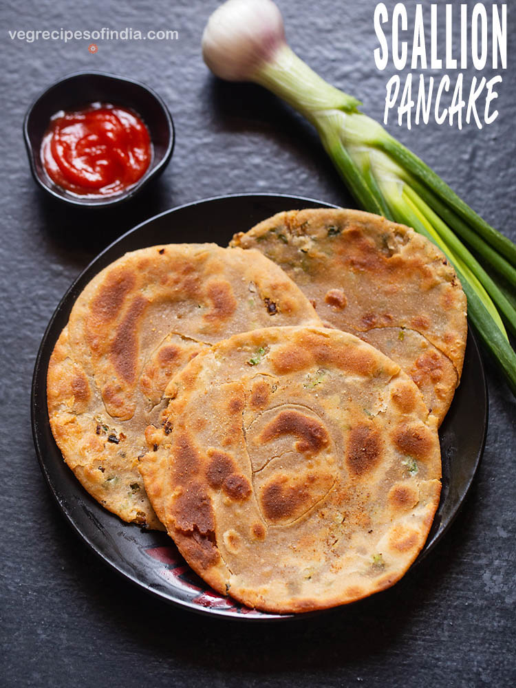 scallion pancakes placed on a black ceramic plate on a black board with sriracha sauce served in a small black bowl on left side and a stalk of spring onion placed on the right side of the plate.