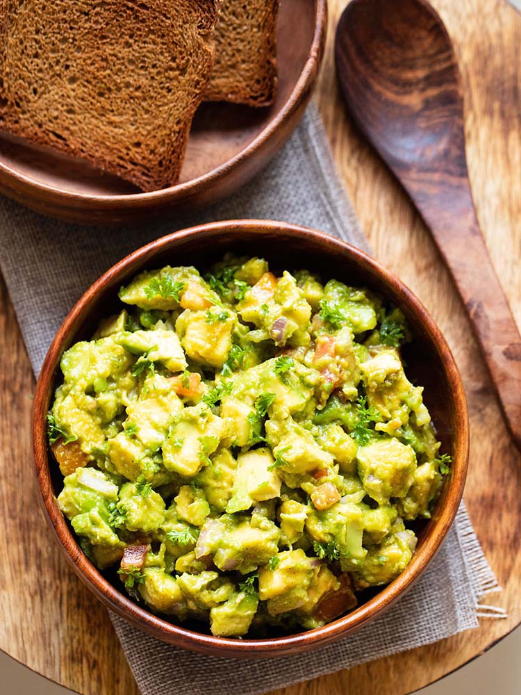 avocado salad served in a wooden bowl with a wooden spoon and some toasted bread in the background