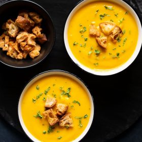 carrot soup served in two cream ceramic bowls on a black round board. topped with croutons and parsley.