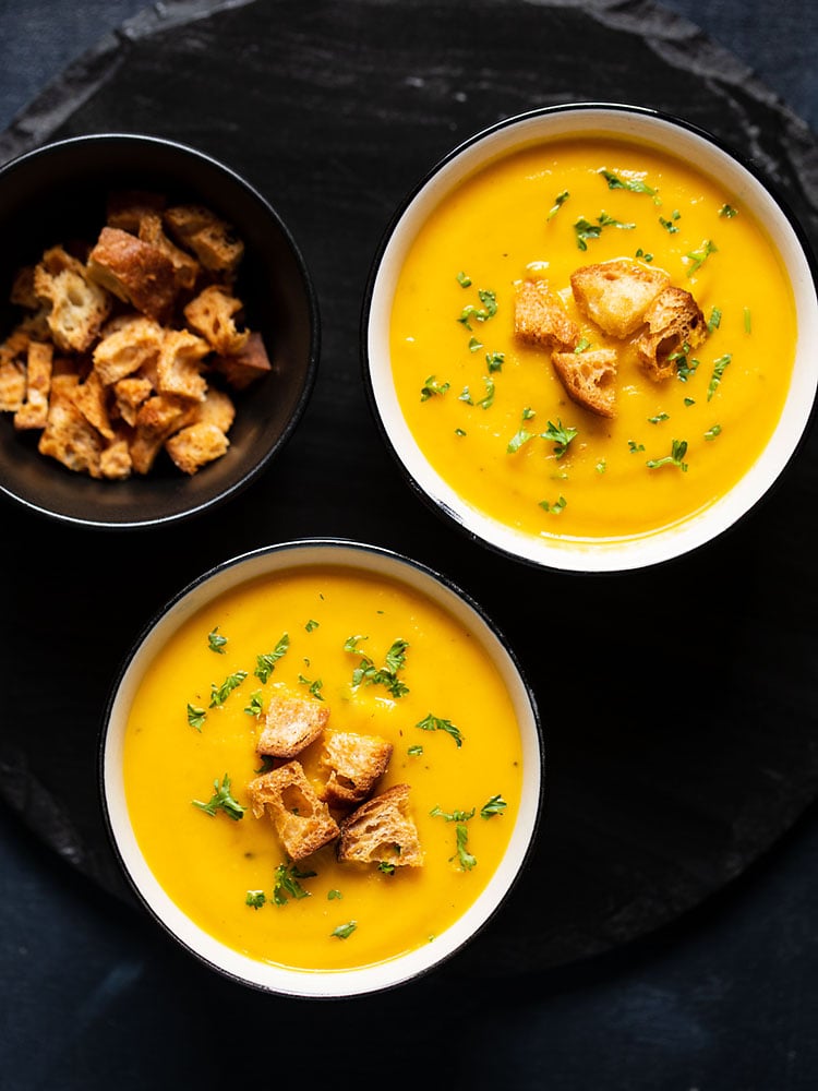 carrot soup served in two cream ceramic bowls on a black round board. topped with croutons and parsley.